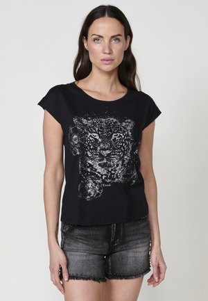 Camiseta de algodón negra con un diseño gráfico de leopardo plateado, mangas cortas y acentos de encaje texturizado a lo largo de las costuras. Combinada con pantalones cortos de mezclilla.