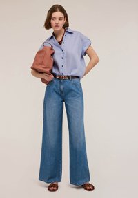 Camisa de botones de manga corta azul claro, pantalones vaqueros de pierna ancha en azul, cinturón marrón y bolso de mano de textura marrón. Lleva sandalias marrones.