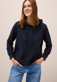 Ung kvinde smilende, iført en marineblå hoodie med snore og lyseblå jeans, hænderne i lommerne, mod en ensfarvet beige baggrund.