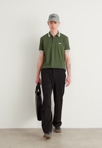 Grønn polo skjorte med hvite detaljer, svarte bukser, grå caps og svart bag. Modellen vist i en nøytral setting, går fremover.