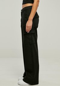 Schwarze Cargo-Hosen mit lockerer Passform, seitlichen Taschen und Knopfverschluss. Der Stoff ist glatt mit einem leichten Glanz; kombiniert mit weißen Sneakern.