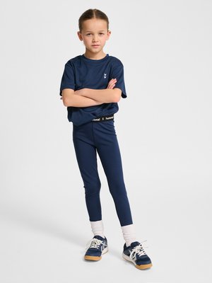 Young girl in navy blue sportswear with arms crossed, standing with white socks and navy sneakers against a plain background.