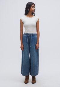 Ελαφρύ κρεμ T-shirt και παντελόνι από μπλε τζιν με φαρδύ πόδι, ελαστική μέση και κορδόνι, φορεμένα με καφέ μποτάκια αστραγάλου.