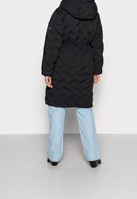 Osoba oblečená v černé kapuci s péřovou bundou a světle modrými širokými džíny stojí na šedé podlaze na pozadí bílé stěny.