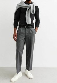 Pantalon à carreaux gris avec plis à l'avant, associé à une chemise noire à manches longues et un pull gris clair drapé sur les épaules. Baskets blanches.
