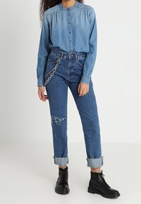 Chemise en denim bleu clair avec fermeture à boutons, assortie à un jean bleu déchiré taille haute avec revers retroussés et bottes noires à lacets.