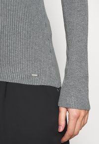 Pull gris côtelé avec un tricot texturé, des manches allongées et un petit logo argenté au bas.