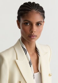 Žena s vrkočmi má na sebe textúrované krémové sako so zlatými gombíkmi a vzorovanú šatku cez biely top, tvárou vpredu.