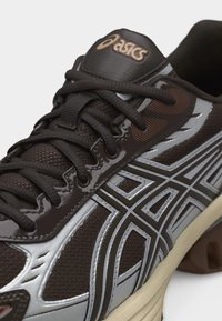 Nahaufnahme eines schwarzen und silbernen ASICS Laufschuhs mit atmungsaktivem Mesh-Obermaterial, schwarzen Schnürsenkeln und beiger Sohle, die das Markenlogo auf der Zunge zeigt.