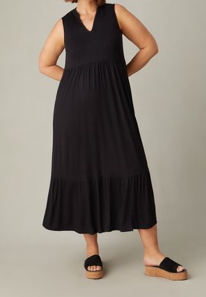 Czarna maxi sukienka bez rękawów z dekoltem w kształcie litery V, posiada zebrany pasek w talii oraz warstwowy krój spódnicy. Nosi ją w połączeniu z czarnymi sandałami na koturnie.