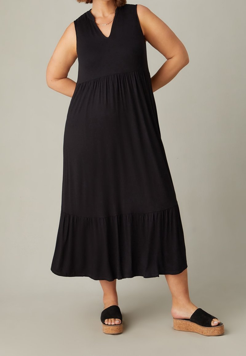 Schwarzes, ärmelloses Maxikleid mit einem V-Ausschnitt, das eine geraffte Taille und ein gestuftes Rockdesign aufweist. Trägt schwarze Plateausandalen.