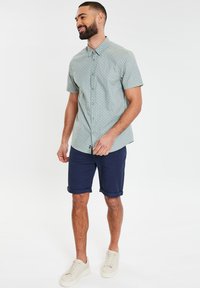 Kurzärmeliges Hemd in Hellblau mit geometrischen Mustern, kombiniert mit navyblauen Shorts und cremafarbigen Sneakers.