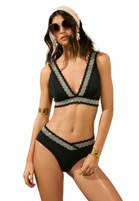 Fekete bikiniszett, v-nyakú bralette-tel csipke díszítéssel és hozzáillő magas derekú alsóval, kontrasztos arany kiegészítőkkel kiemelve.