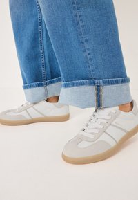 Sneaker bianchi e grigi in pelle scamosciata e pelle liscia, con suole in gomma e chiusura con lacci. Indossati con jeans di denim blu arrotolati.