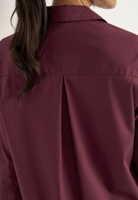 Rotes Hemd mit einer Rückfalte, aus glattem Stoff gefertigt. Verfügt über einen klassischen Kragen und einen lässigen Schnitt, der eine tiefe burgunderrote Farbe zur Geltung bringt.