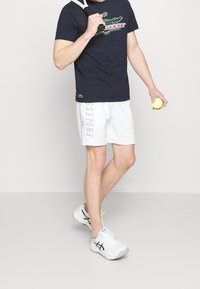 Navyblå t-shirt med ett krokodiltryck, matchad med vita shorts med texten "ENDLESS". Vita sportskor och ett tennisracket.