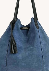 Sac fourre-tout en daim bleu avec des poignées en cuir noir et un pompon, arborant une petite étiquette dorée indiquant "Loxwood Le Cabas Parisien."