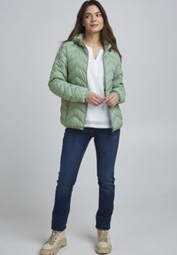 Veste rembourrée matelassée vert clair avec capuche, associée à un haut blanc et un jean bleu foncé. Des bottes beige à lacets complètent la tenue.