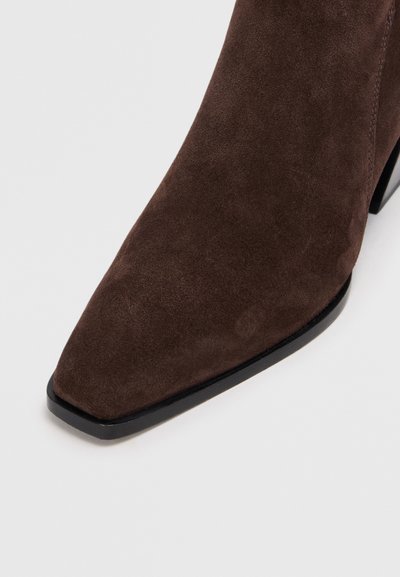 Bottine en suède marron avec un bout pointu, présentant une texture lisse et un talon noir empilé pour plus de stabilité.