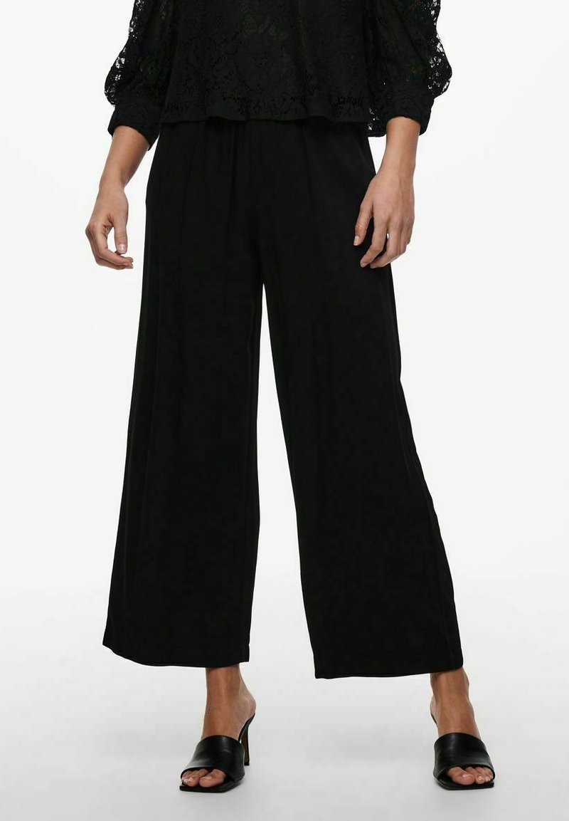 Femme portant un pantalon noir à jambes larges, un haut en dentelle noire, et des mules à talons ouvertes noires, debout sur un fond blanc.