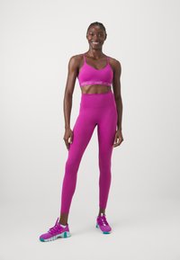 Fuchsiová sportovní podprsenka s logem Nike, odpovídající fuchsiové legíny s vysokým pasem a purpurové sportovní boty s akcenty v barvě tyrkysu. Hladká textura.