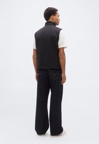 Ανδρας που φοράει μαύρο αμάνικο γιλέκο πάνω από λευκή μπλούζα, σκούρο τζιν με φαρδύ μπατζάκι και λευκά αθλητικά παπούτσια, στραμμένος προς τα πίσω μπροστά σε απλό φόντο.