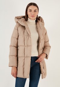 Béžová péřová bunda s širokou kapucí, prošívaným designem a střední délkou. Nosená přes krémový rolák a v kombinaci s tmavými džíny.