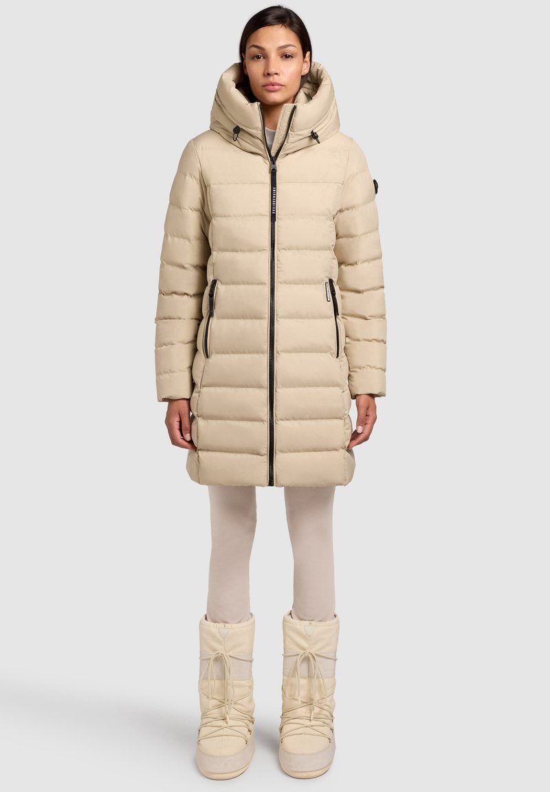Beige dunjakke med høj krave, frontlynlås og to sidelommer. Sammen med lyse støvler og leggings for et sammenhængende look.