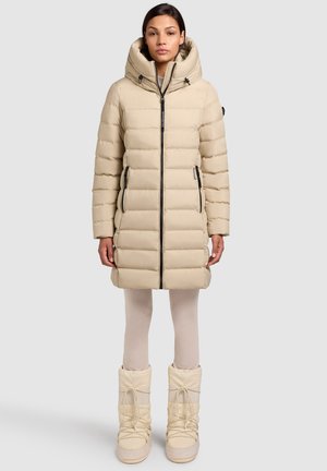 Beige puffer coat with a high collar, front zipper, and two side pockets. Paired with light-colored boots and leggings for a cohesive look.