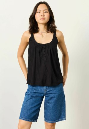 Tânără purtând o bluză neagră fără mâneci și pantaloni scurți din denim albastru, stând cu mâinile în buzunare pe un fundal deschis.