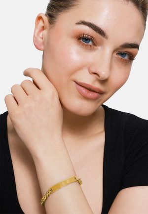 Bracciale in tonalità oro con una piastra centrale piatta e liscia, caratterizzata da un design a catena. Indossato su un polso di carnagione chiara, con una maglia nera.