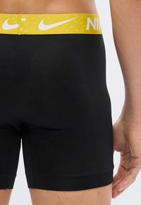 Άνδρας που φοράει μαύρα μποξεράκια με κίτρινη μέση διακοσμημένη με λευκά λογότυπα Nike, εμφανιζόμενος από τη μέση και κάτω, με το δεξί χέρι ορατό.