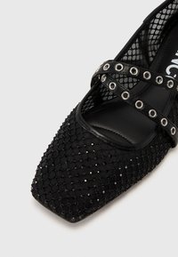 Chaussure plate noire avec un dessus en maille texturée et un bout carré, ornée d'accents cloutés et de sangles croisées.