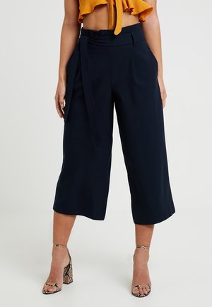 Navy culottes with a high waist, front pleats, and a tie belt. Smooth fabric with a wide-leg cut, paired with strappy, patterned heels.