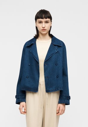 Młoda kobieta o krótkich ciemnych włosach nosi ciemnoniebieską krótką kurtkę jeansową na beżowej bluzce i jasnobeżowych spodniach, stojąc na jednolitym tle.