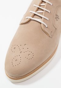 Chaussure en suede beige avec un bout arrondi, présentant des détails perforés et des lacets blancs. Semelle blanche avec des accents marron clair.