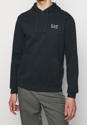 Felpa nera con cappuccio e tasca frontale, con logo bianco "EA7 Emporio Armani" sul petto a sinistra, indossata con pantaloni grigi.