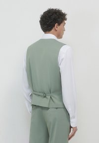 Grüne maßgeschneiderte Weste mit passgenauem Design, versehen mit einem Gürtel mit Metallring und Faltendetails auf der Rückseite, getragen über einem weißen Hemd.