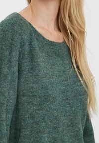 Pull en laine verte avec un large col et une texture douce. Il présente une coupe ajustée et une légère aisance au niveau des épaules.