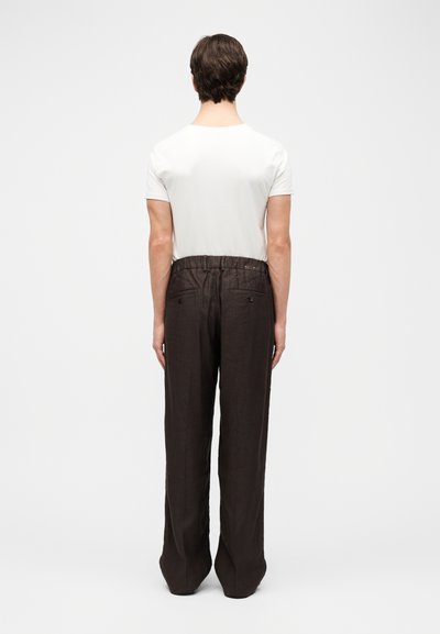 Άνδρας που στέκεται με την πλάτη, φοράει λευκό κοντομάνικο πουκάμισο και άνετο σκούρο καφέ παντελόνι με πίσω τσέπες.