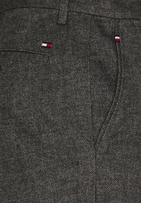 Donkergrijze broek van textuurstof, met een klein driekleurig logo (rood, wit, blauw) op de zak en discrete stiksels.