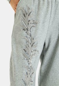 Graue Sweatpants mit einem dekorativen silbernen Paillettenblattdesign an der Seite, aus glattem Stoff und mit seitlichen Taschen.