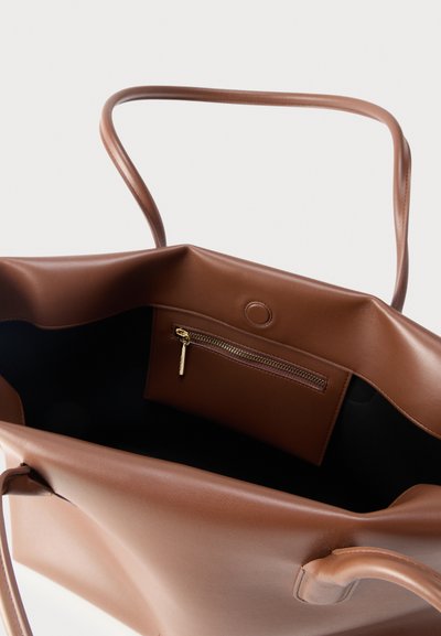 Καφέ τσάντα ώμου από δέρμα με μαλακή δομή, ανοιχτό πάνω μέρος, εσωτερική τσέπη με φερμουάρ και λεπτούς ιμάντες ώμου σε απλό φόντο.