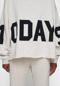 Sudadera gris claro con texto negro "100 DÍAS"; presenta un corte holgado, hombros caídos y puños acanalados. Se combina con pantalones a juego.