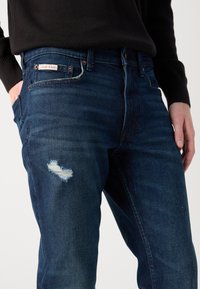 Tamno plave traperice s oštećenim detaljima na koljenu, s kontrastnim šavovima, oznakom Calvin Klein na pojasu i uskim krojem.