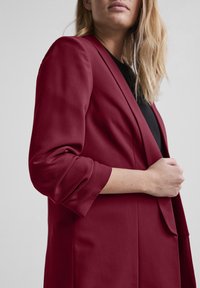 Blazer bordeaux à coupe slim, épaules structurées, manches retroussées et texture lisse. Doté d'une fermeture à un bouton et de revers crantés.
