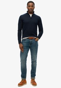 Marine kwart rits trui, witte shirt eronder, lichtblauwe jeans en bruine leren laarzen. Eenvoudig ontwerp met textuurbreiwerk en casual pasvorm.