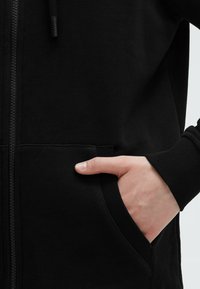 Black zip-up hoodie made of soft fabric with ribbed cuffs and pockets. The zipper is along the center front. Smooth texture.