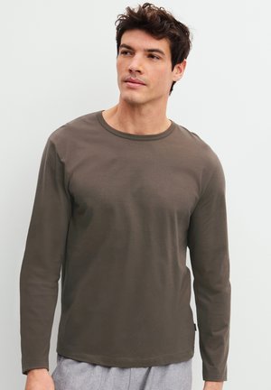 Jonge man met donker haar die een olijfgroen overhemd met lange mouwen en lichtgrijze broek draagt, staand tegen een effen lichte achtergrond.