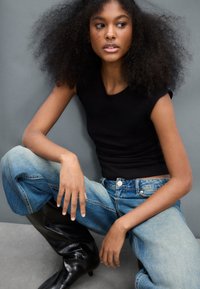 Musta istuva hihaton toppi, vaaleansiniset korkeavyötäröiset farkut, joissa haalistunut viimeistely, ja polven korkuiset mustat kiiltävät saappaat. Harmaa tausta.